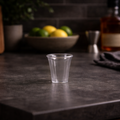 Clear shot glass on a dark countertop with a blurred background of fruits and a bottle.
